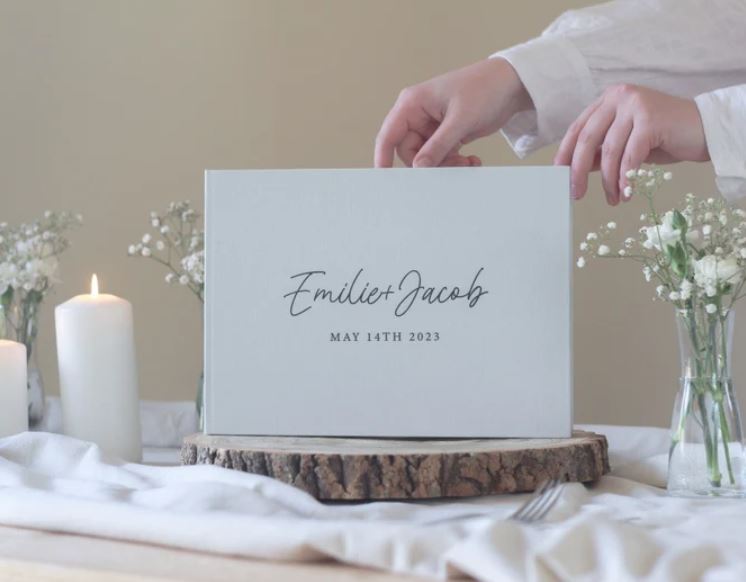 White hardcover wedding guest book personalized with Emilie and Jacob May 14th 2023 displayed on a wood slice with baby's breath and a white pillar candle