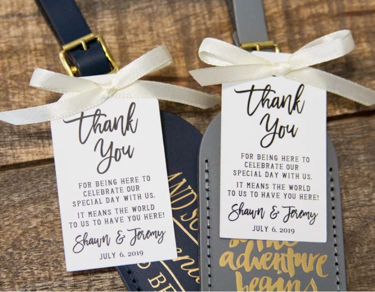 Navy and gray leather luggage tags tied with ivory ribbon bows and personalized thank-you cards for Shawn and Jeremy displayed on a wood surface