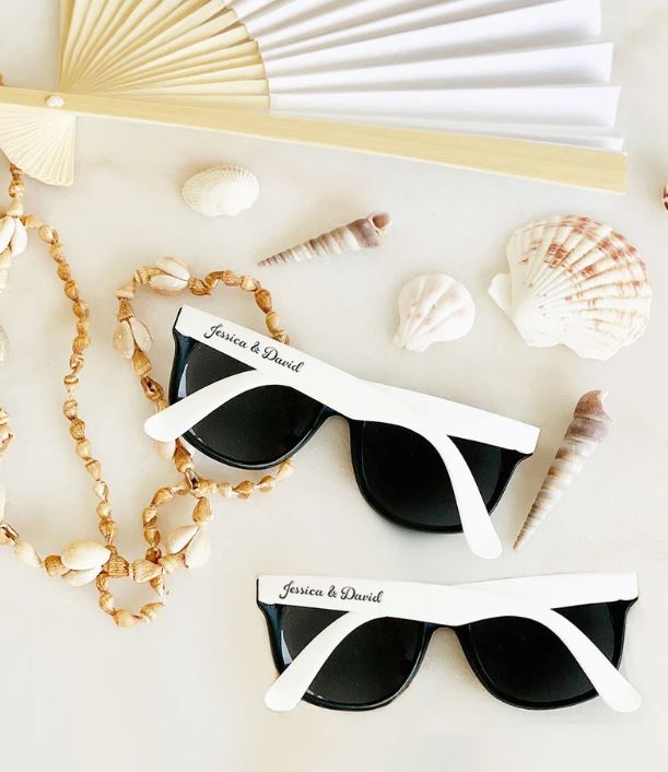 Two pairs of black and white sunglasses personalized with Jessica and David displayed among seashells and a hand fan on a white surface
