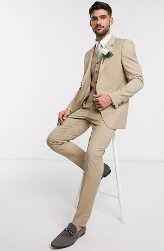 Man in a tan three-piece slim wedding suit with a white rose boutonniere and grey loafers, seated on a stool in a studio product photo