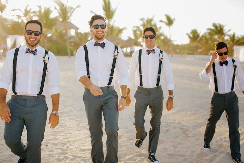 Four groomsmen in grey dress pants, white shirts, black suspenders, and patterned bowties wearing sunglasses and walking on a sandy beach at a tropical resort