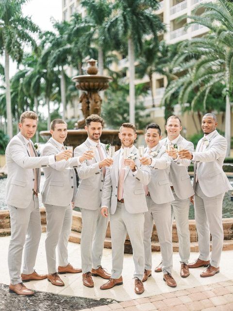 Seven groomsmen in matching light grey suits with pink ties toasting champagne glasses in front of a resort fountain surrounded by palm trees