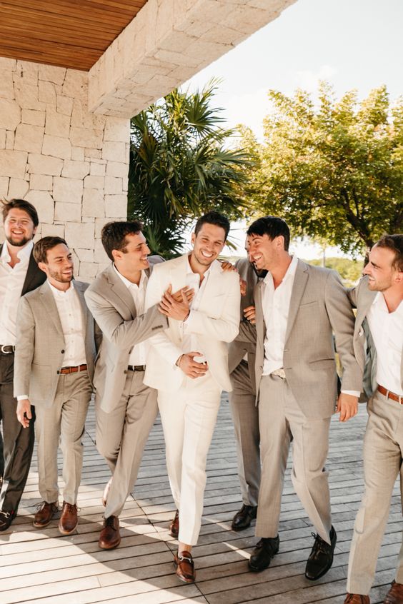 Groom in a cream suit laughing and celebrating with six groomsmen in light grey suits at a tropical resort with palm trees