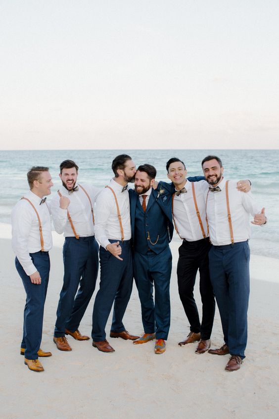Groom in a navy three-piece suit with groomsmen in navy pants, white shirts, and brown leather suspenders celebrating together on a beach at sunset