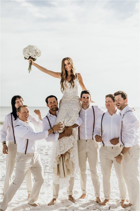 Groomsmen in white shirts and brown suspenders lifting a laughing bride in a white fitted lace gown high above the sand on a beach