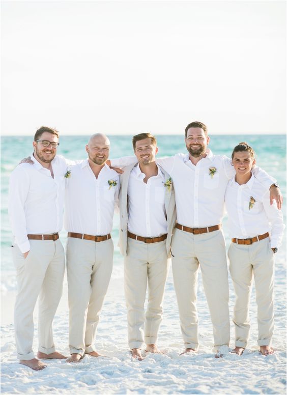 Five barefoot groomsmen in white dress shirts and tan linen pants laughing together on a white sand beach with turquoise water