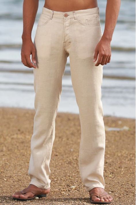 Man wearing natural beige relaxed-fit linen pants with sandals, standing on a sandy beach with the ocean in the background