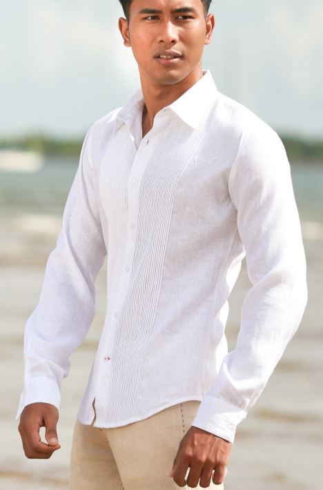 Man wearing a white long-sleeve linen shirt with pintuck pleating down the front, standing on a beach with the ocean behind him