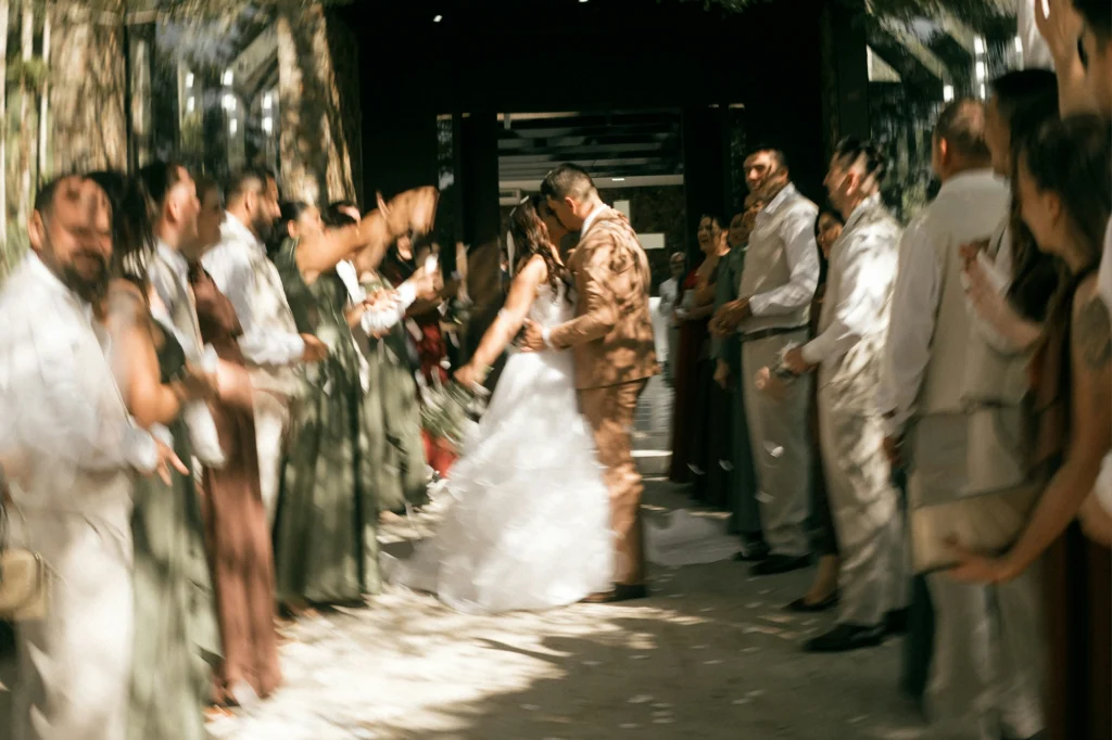 A newlywed couple walks hand in hand through a crowd of wedding guests outside a building, with strong motion blur creating a lively, celebratory effect.