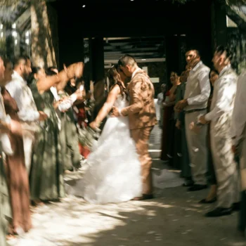 A newlywed couple walks hand in hand through a crowd of wedding guests outside a building, with strong motion blur creating a lively, celebratory effect.