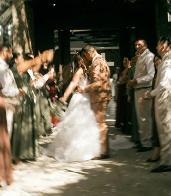 A newlywed couple walks hand in hand through a crowd of wedding guests outside a building, with strong motion blur creating a lively, celebratory effect.