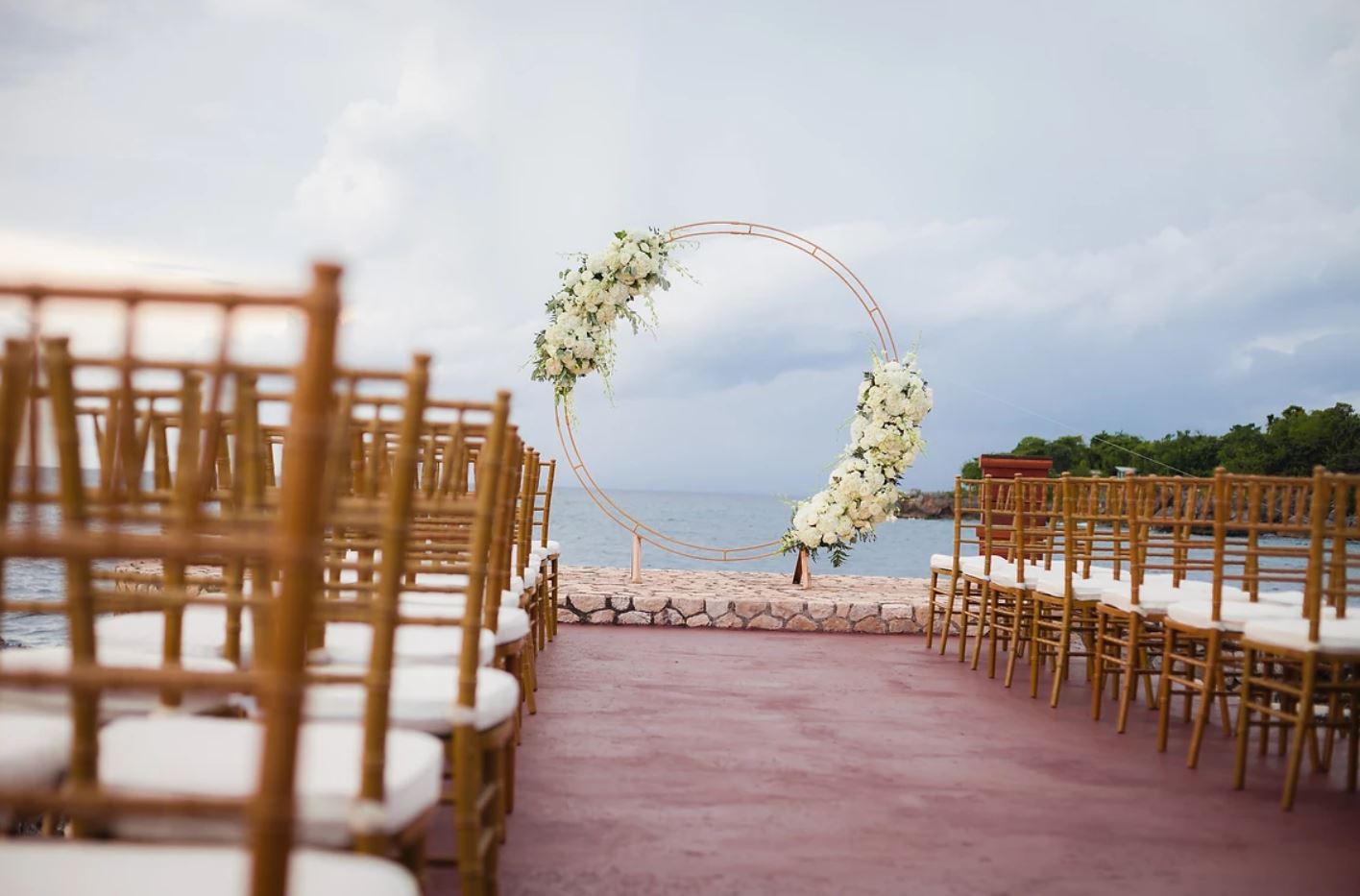 Outdoor destination wedding ceremony setup with rows of gold chiavari chairs and a rose gold circular arch covered in white roses overlooking the water