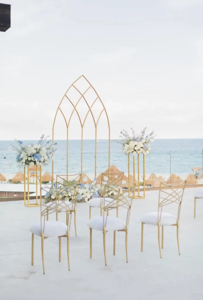 Beachfront destination wedding ceremony setup with gold chairs, floral arrangements, and modern gold arch—featured by Breezy Brides.