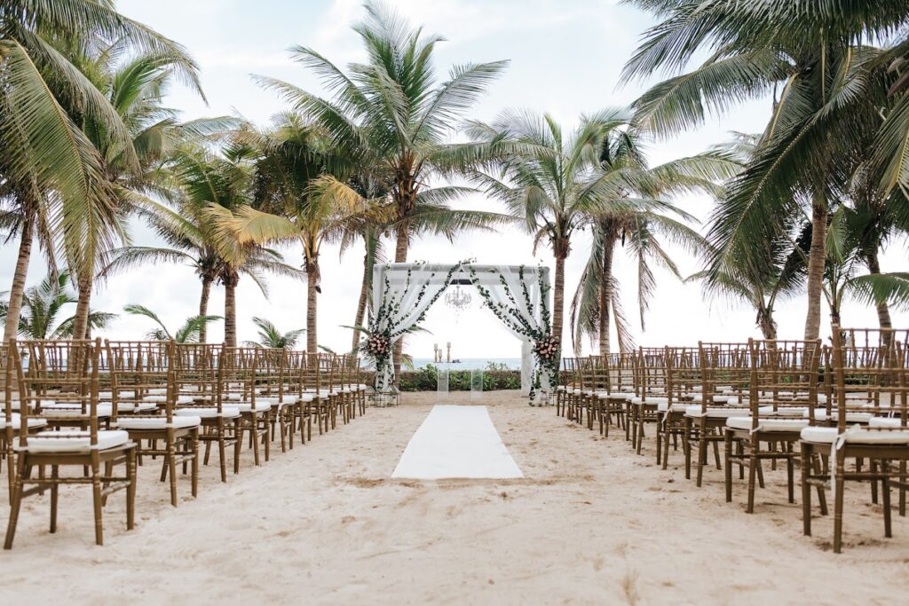 Beachfront wedding ceremony at El Dorado Resort: a draped altar with chandelier and greenery, rows of gold chairs, framed by palm trees and ocean.
