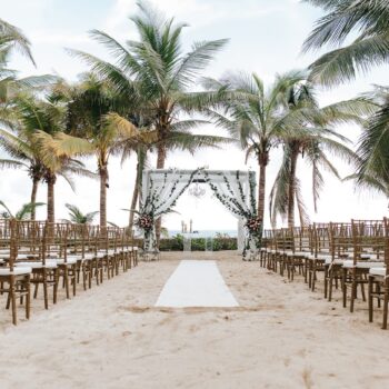 Beachfront wedding ceremony at El Dorado Resort: a draped altar with chandelier and greenery, rows of gold chairs, framed by palm trees and ocean.