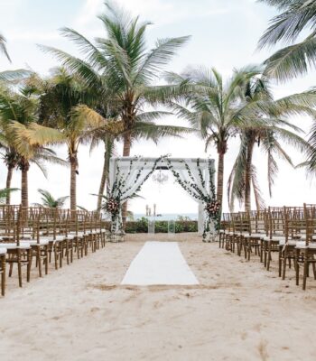 Beachfront wedding ceremony at El Dorado Resort: a draped altar with chandelier and greenery, rows of gold chairs, framed by palm trees and ocean.