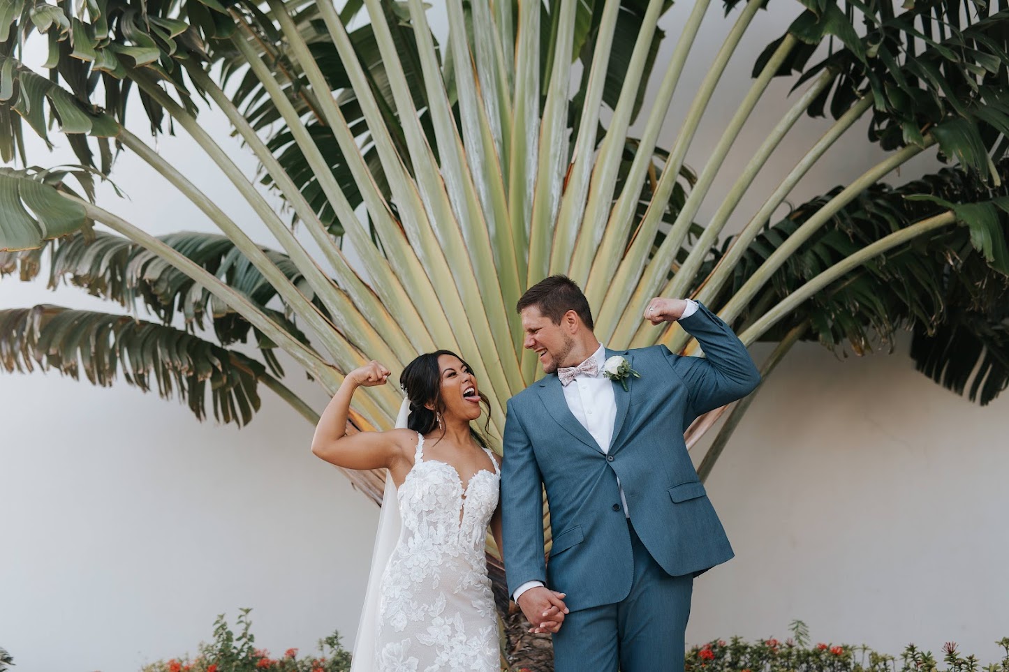Bride in a lace gown and veil and groom in a blue suit laugh while flexing their arms in front of a large fan-shaped traveler's palm.