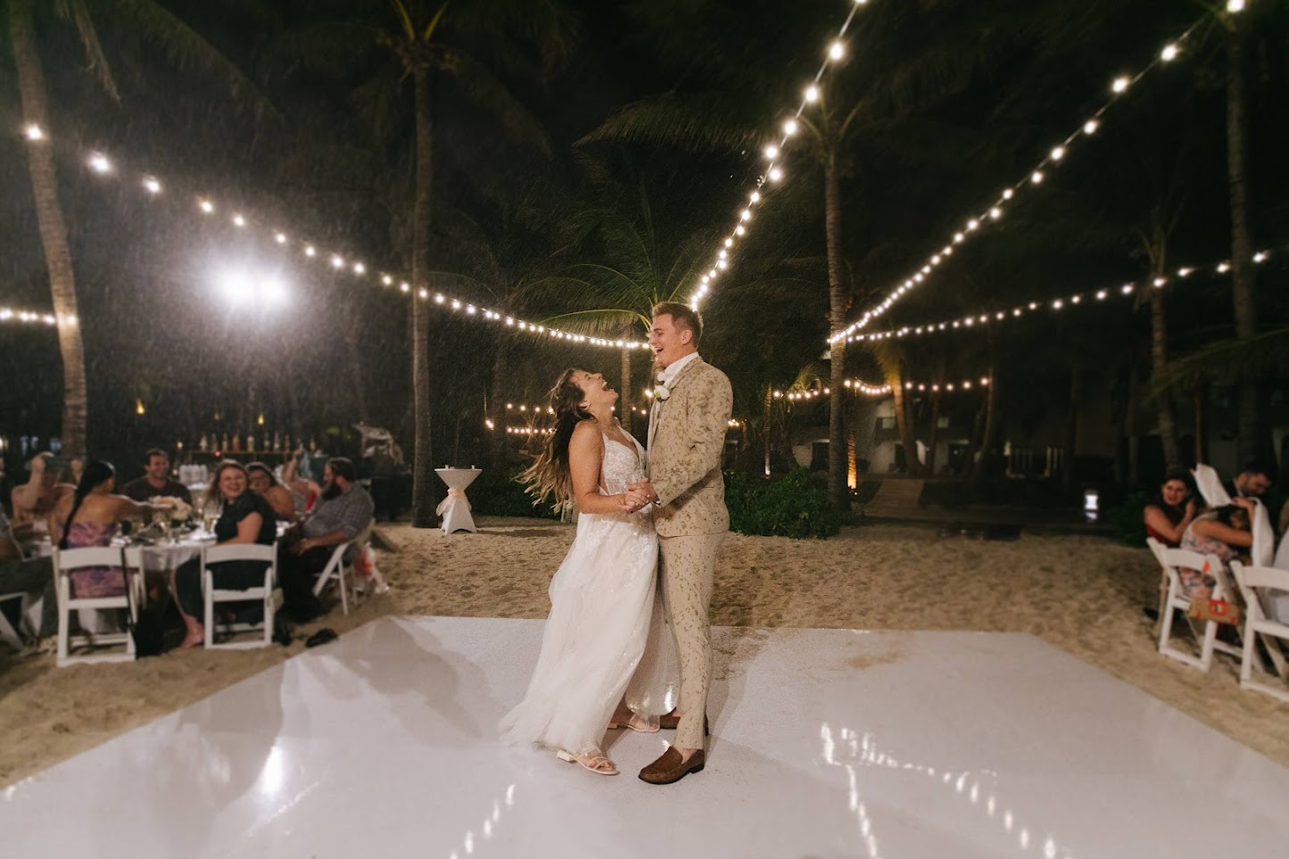 Bride and groom laughing during their first dance on a white beachside dance floor at night, lit by string lights strung between palm trees as seated guests watch.