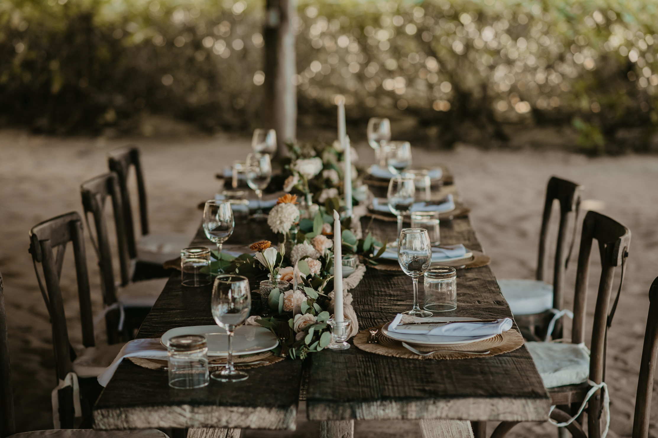 Rustic wooden reception table set on the sand for a wedding in Cancun, with a eucalyptus-and-rose runner, white taper candles, wine glasses, and dark wood chairs.